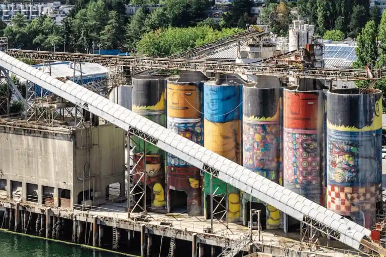 Silos industriais coloridos com murais de arte urbana retratando personagens gigantes e formas abstratas, à beira de um porto cercado por vegetação