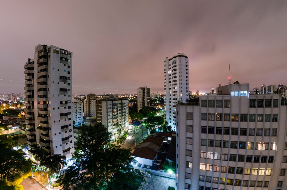 Visão noturna de edifícios residenciais na cidade de São Paulo, iluminados com luzes azuis, verdes e brancas, sob céu nublado.