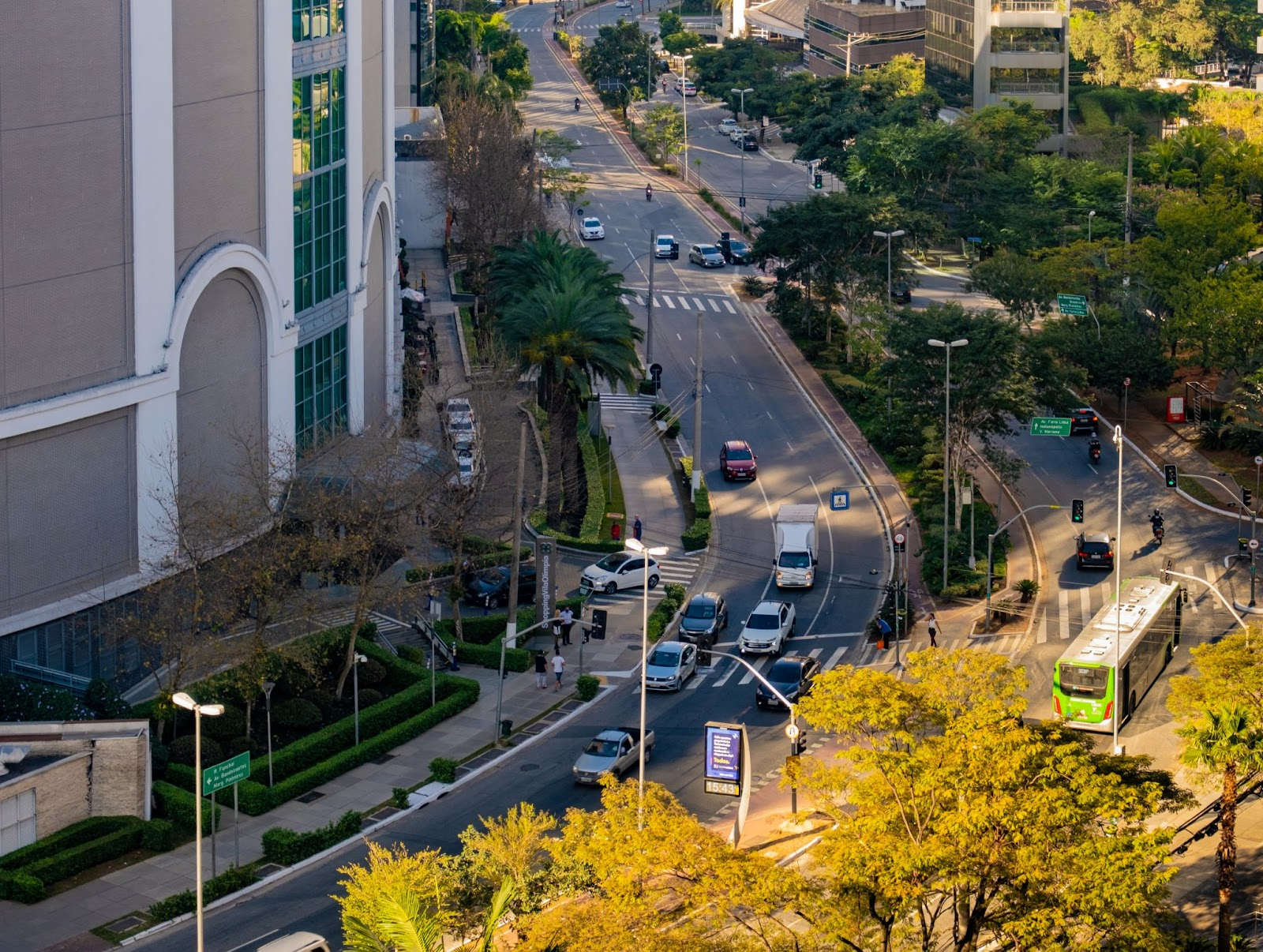Vista aérea de uma rua movimentada em uma cidade moderna com edifícios altos, árvores e veículos, incluindo ônibus e carros, durante o dia.