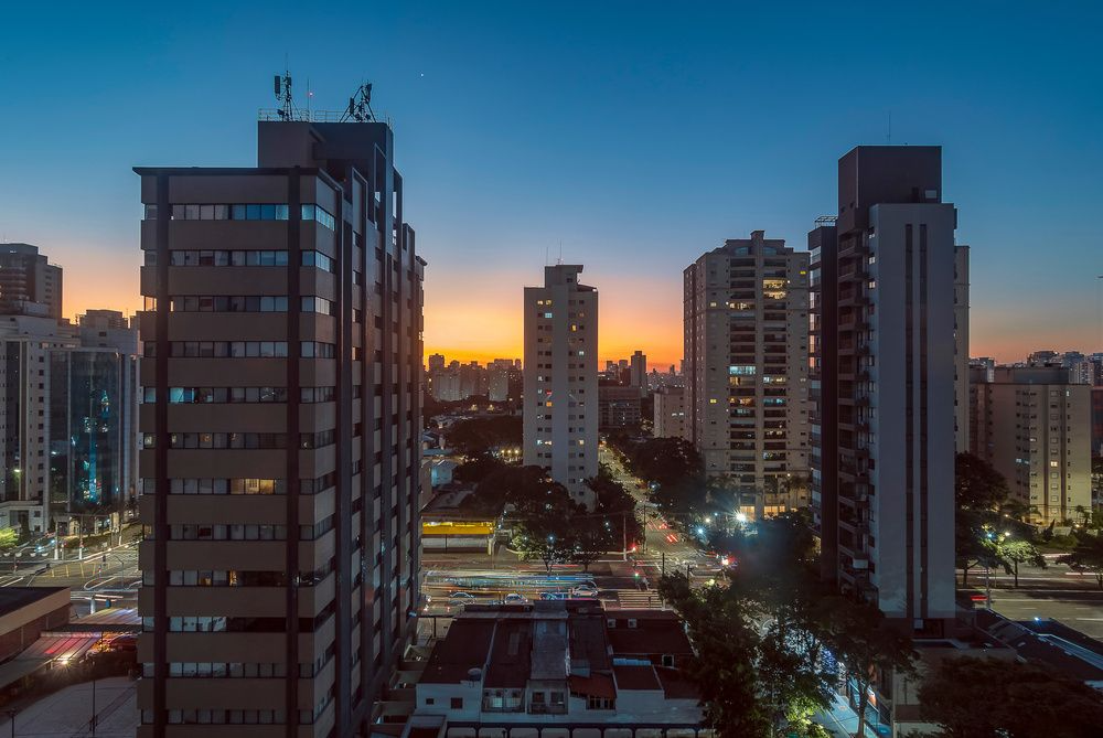 Vista de cidade com prédios altos ao entardecer, com o céu exibindo cores quentes do pôr do sol e movimentação nas ruas.