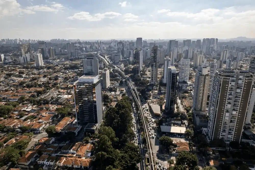 Imagem aérea de Campo Belo, São Paulo, com muitos arranha-céus, avenidas movimentadas e áreas verdes, sob um céu parcialmente nublado.