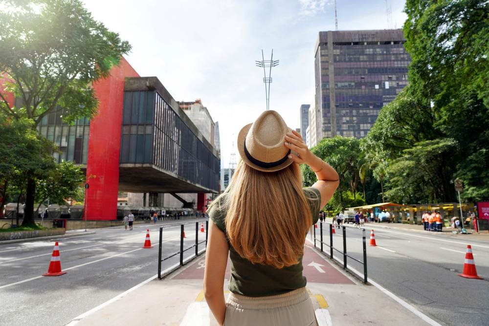 Mulher com cabelo longo usando chapéu, observando o cenário urbano com prédios altos e árvores na Avenida Paulista, em São Paulo.